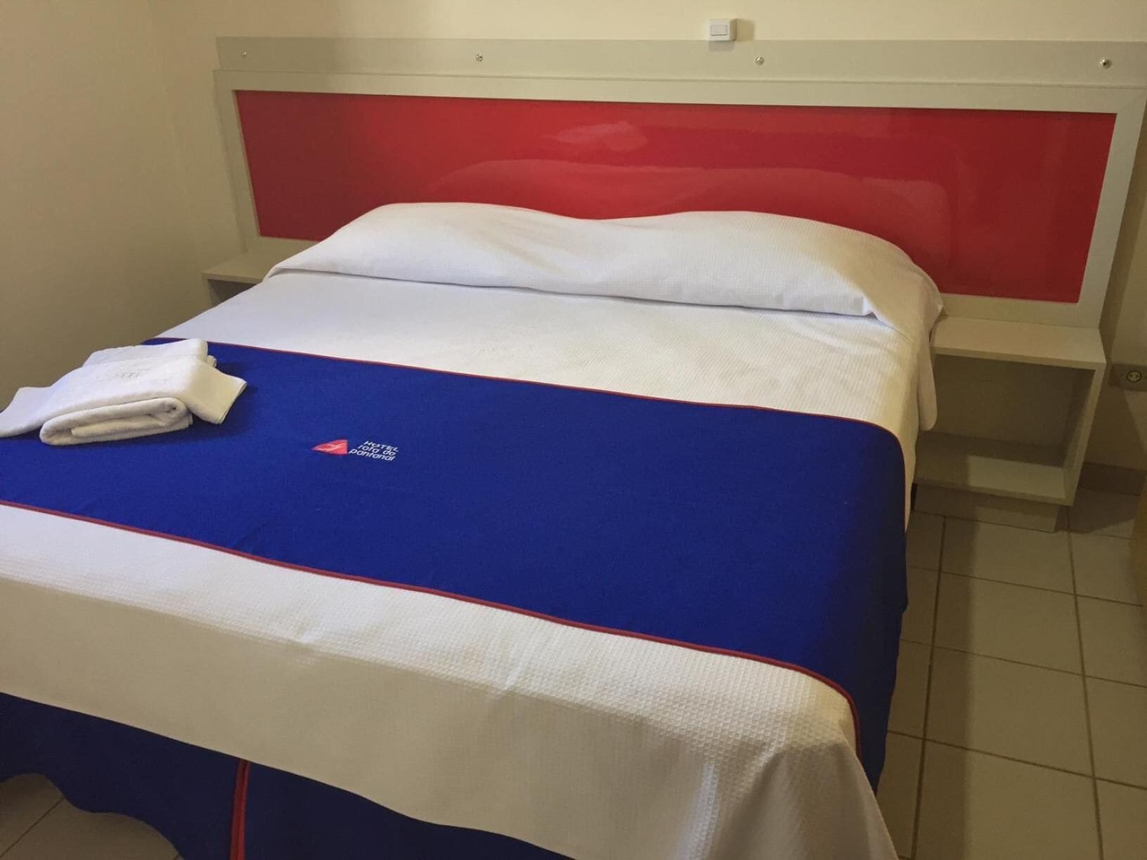 A single bed with a red headboard, a white pillow, and a blue bedspread with a logo.