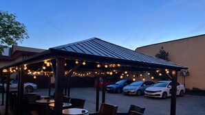 An outdoor seating area with tables and chairs under a covered pavilion, connected to a parking area with several cars. - Hotel Ambassador (Timisoara)