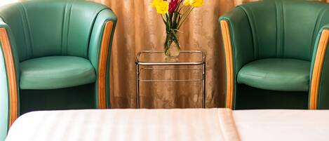 A hotel room with two green upholstered chairs, a small table with a vase of flowers, and a bed with a striped bedspread.