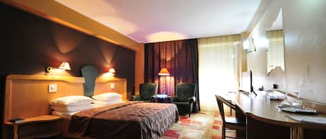 A hotel room with a bed, a desk, two chairs, a lamp, and a window with curtains.