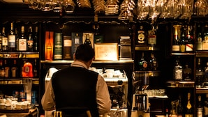 A bar with a bartender preparing drinks, shelves of liquor bottles, and a coffee machine. - Hotel Ambassador (Timisoara)