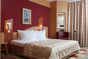 Habitación de hotel con una cama grande, dos lámparas de noche, una pared roja, un espejo, una silla roja y una mesita con un florero.
