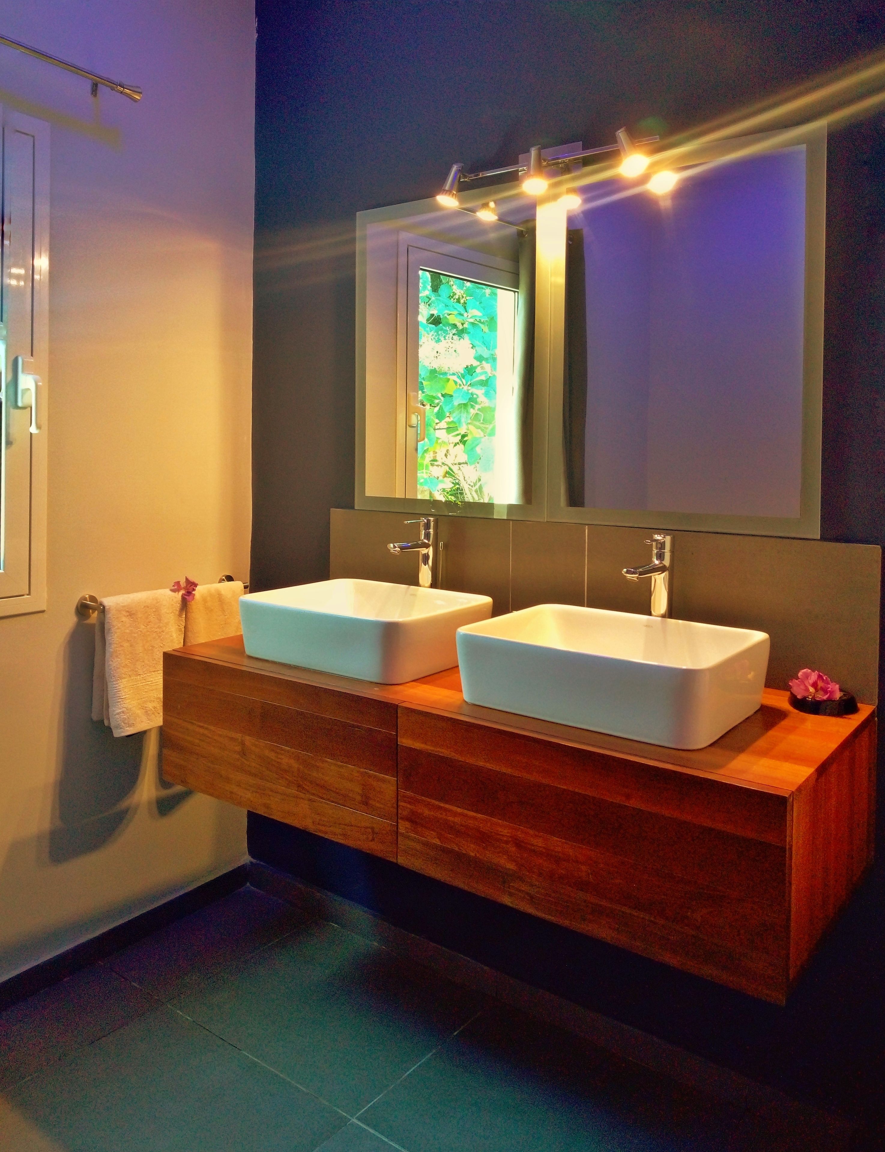 Un baño moderno con dos lavamanos blancos, un mueble de tocador de madera, un espejo grande y una ventana con vistas a la vegetación.