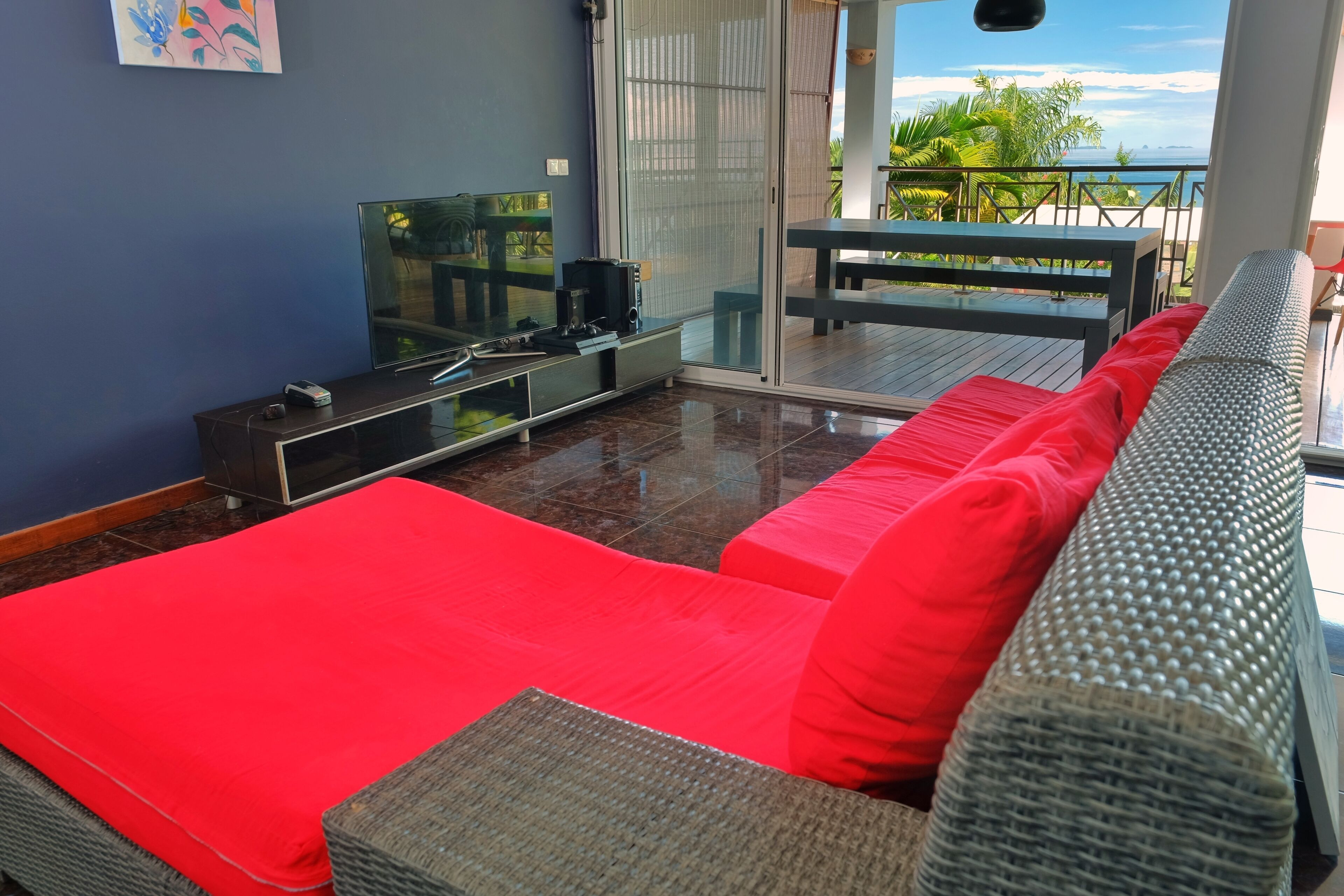 A modern living room with a red sofa, a flat-screen TV, and a glass door leading to a balcony with a view of the ocean.