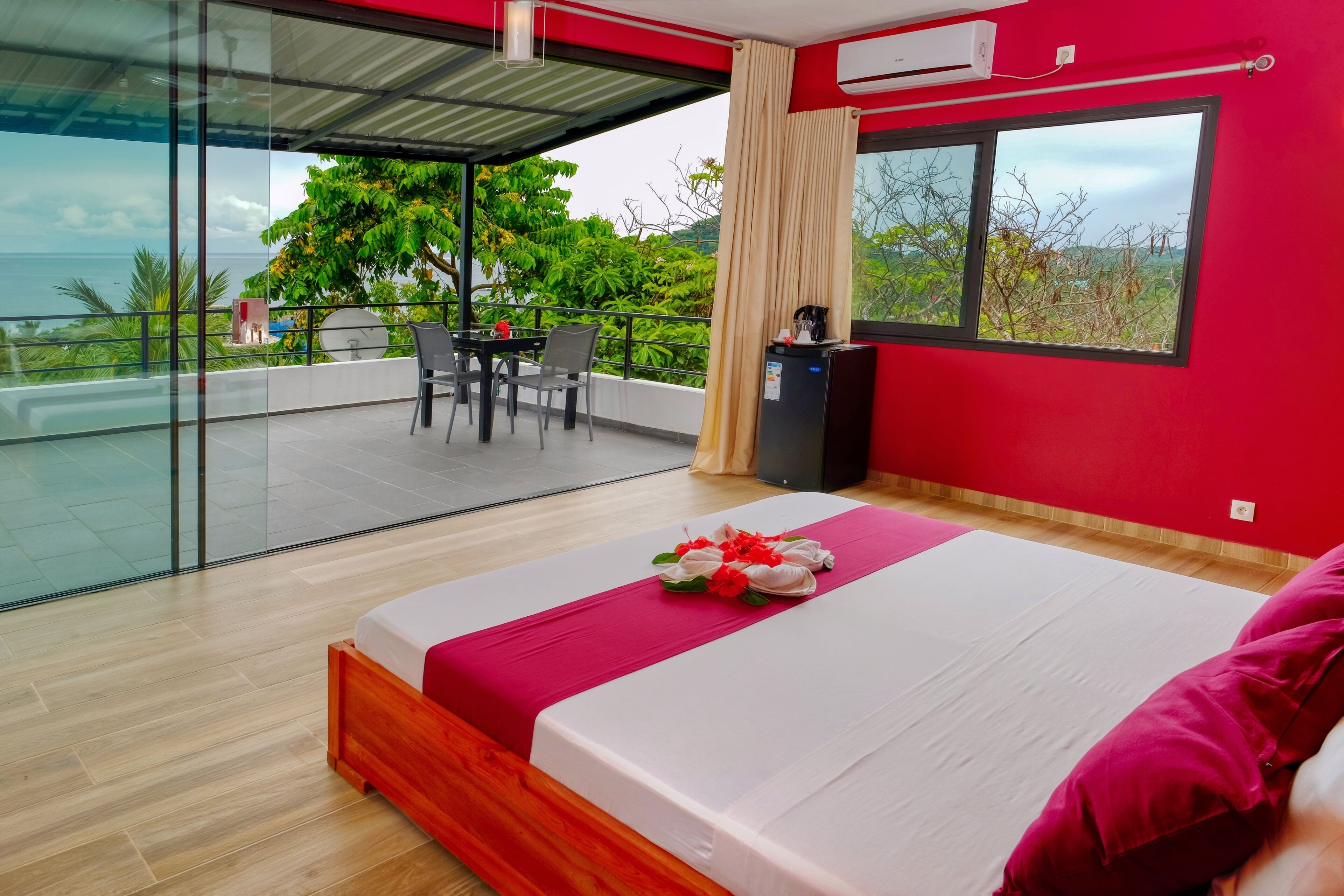 A modern hotel room with a large bed, a balcony with a table and chairs, and a view of the sea.