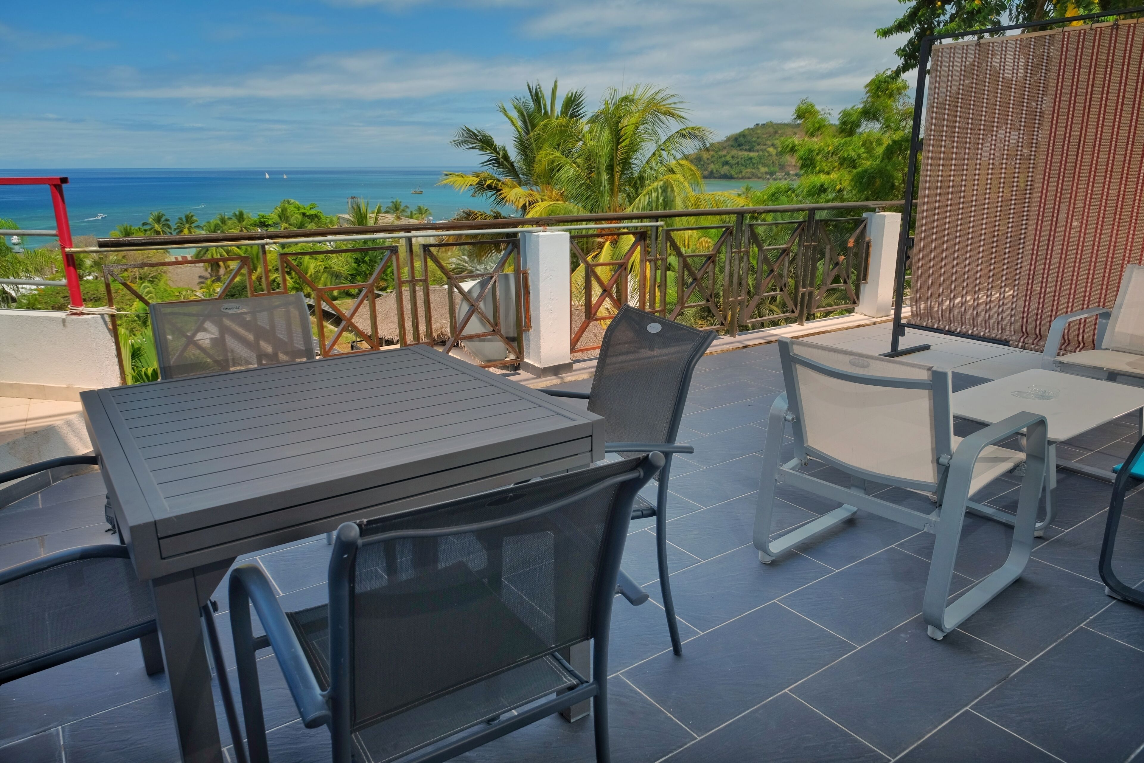 A terrace with a table and chairs, overlooking a coastal view with palm trees and the sea.