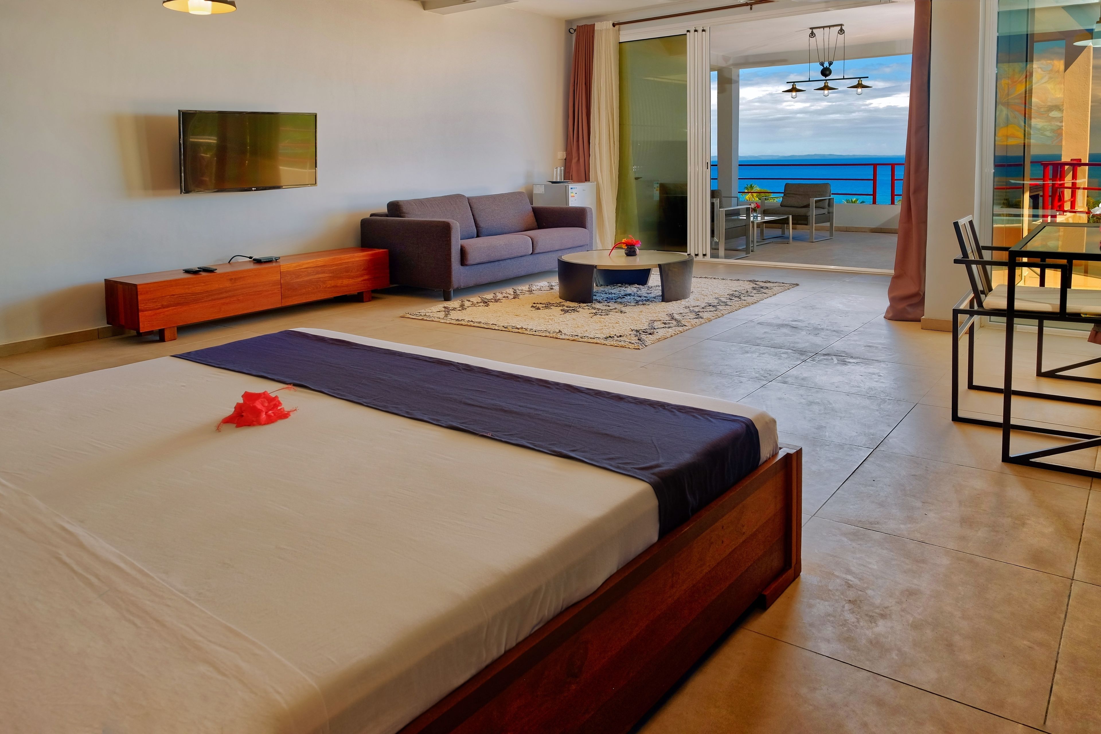 A modern hotel room with a large bed, a sofa, a TV, a small table, and a balcony with a view of the sea.