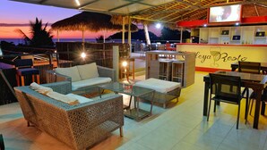 Un rincón acogedor al aire libre con muebles de mimbre y un bar llamado “Calleya Resto” con el atardecer de fondo.