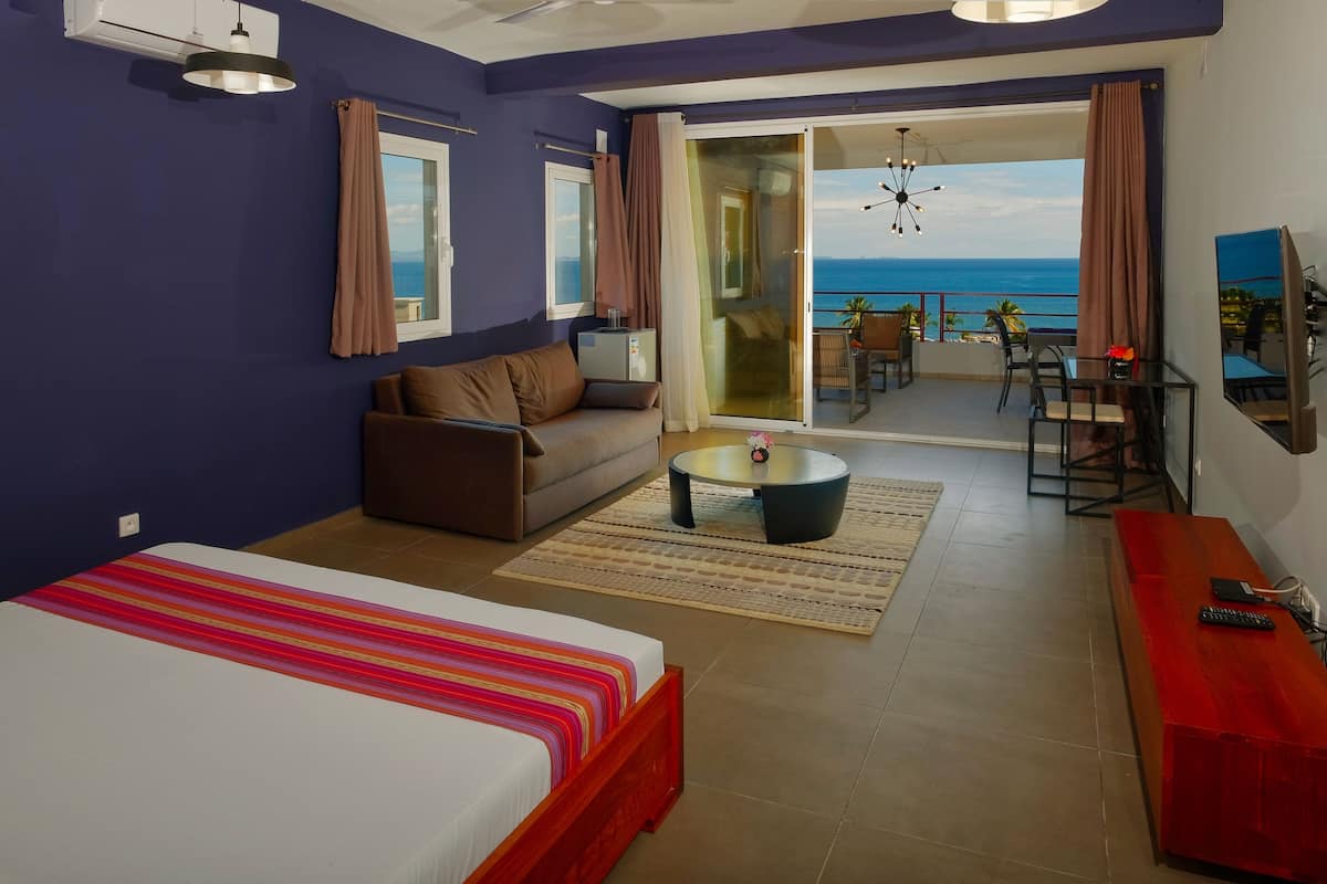 Habitación de hotel con cama, sofá, mesa de centro, televisor y balcón con vista al mar.