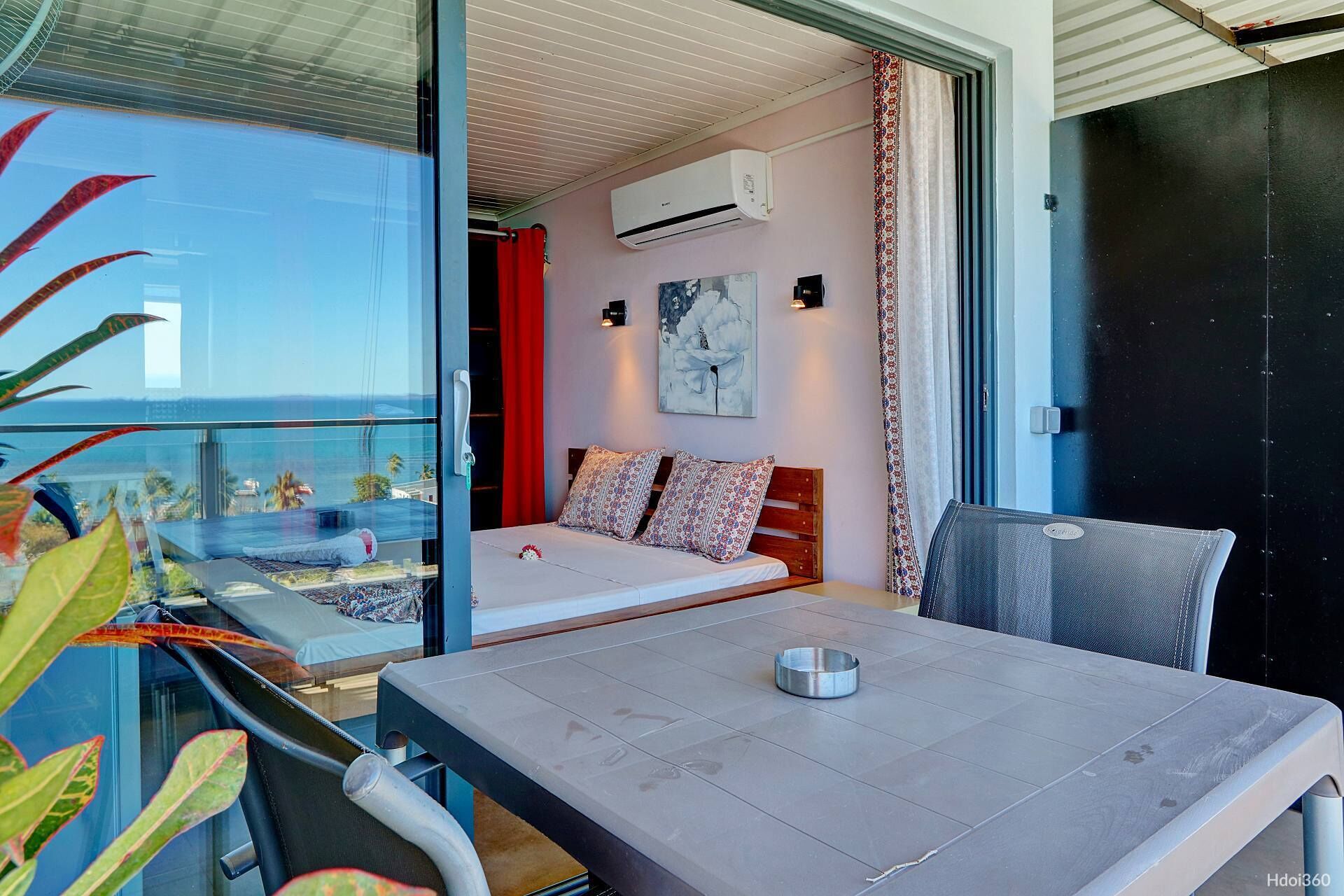 A balcony with a bed, a table, and a chair. There is an air conditioning unit on the wall and a painting above the bed.