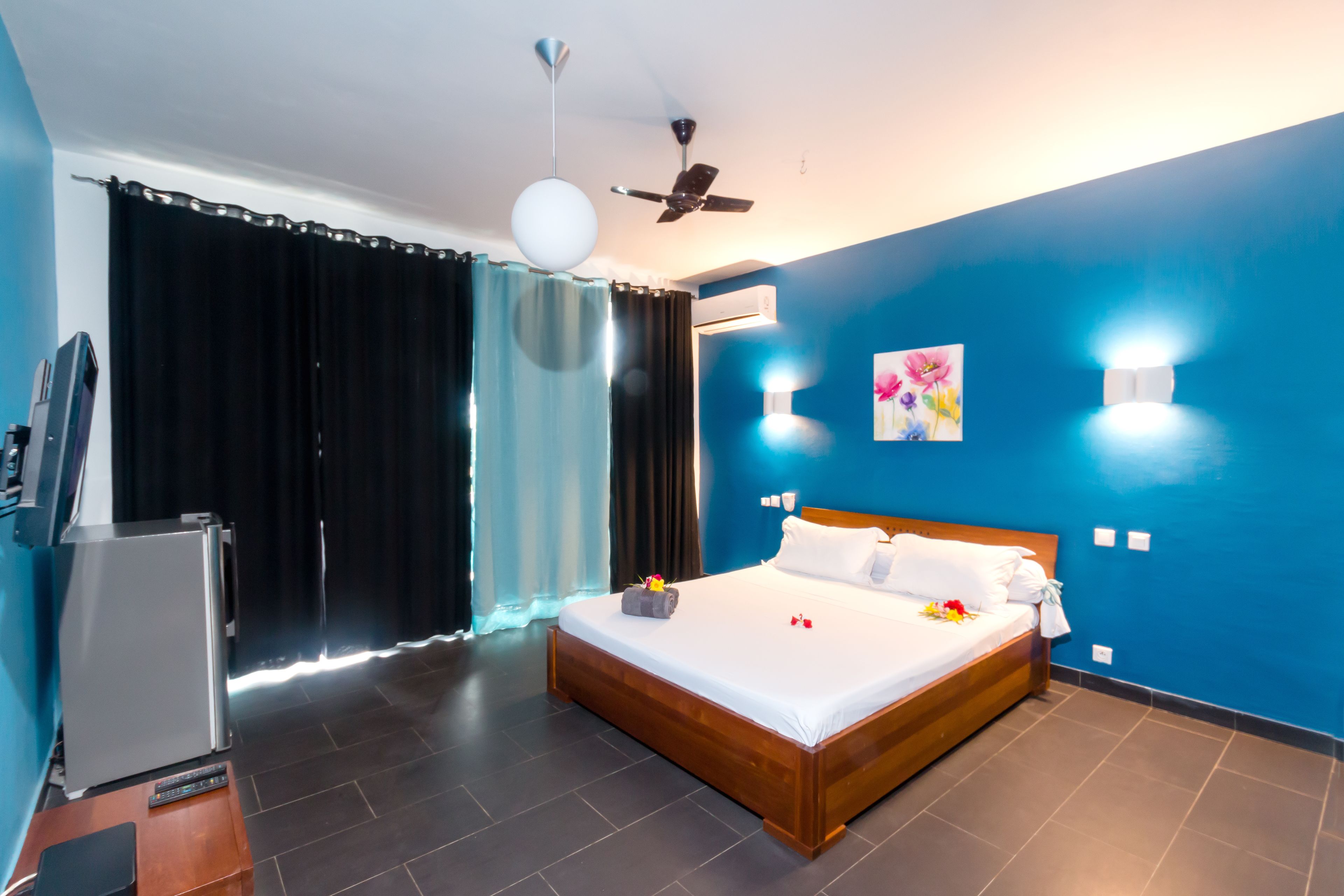 A modern hotel room with a large bed, a ceiling fan, a TV, and a refrigerator.