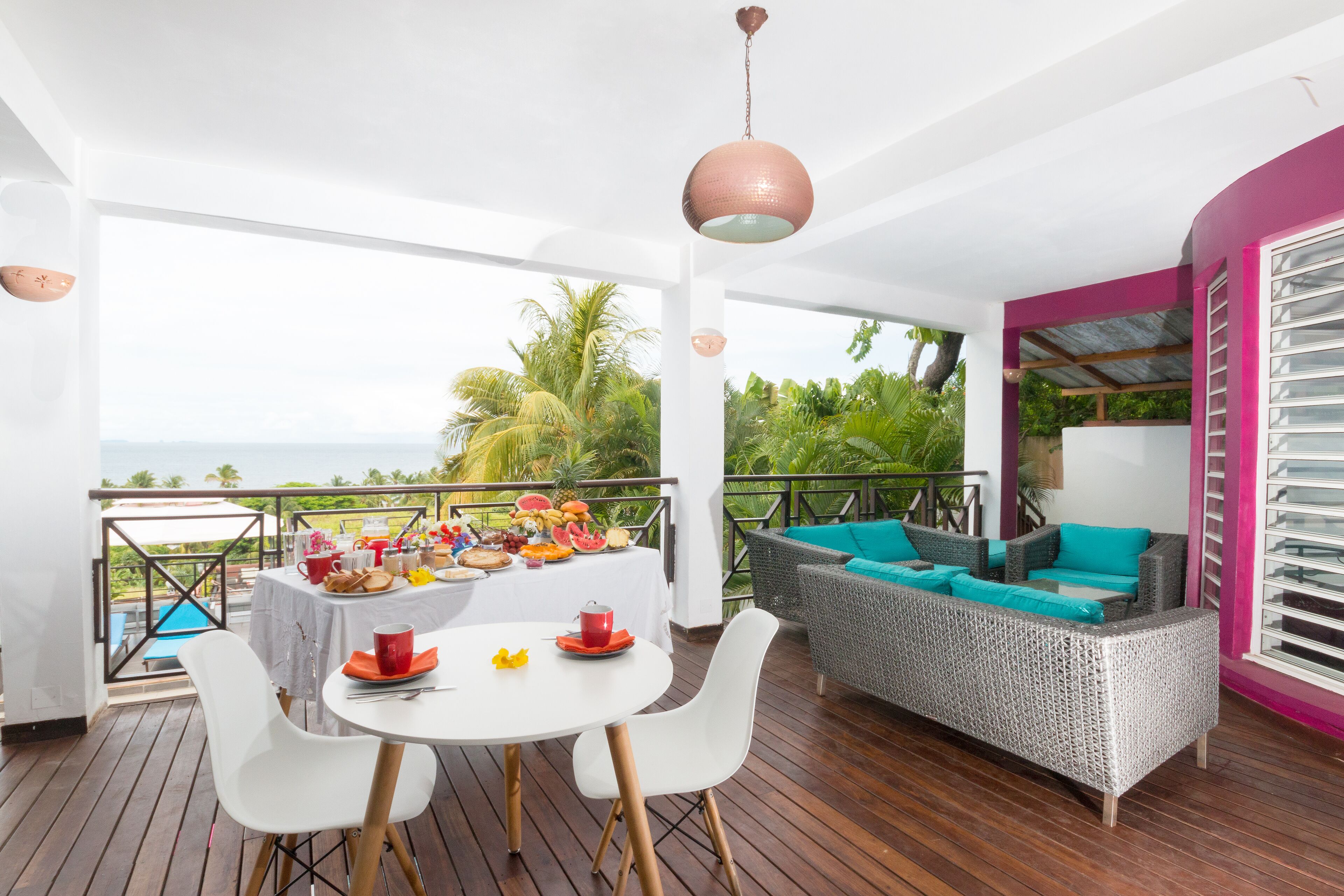 A balcony with a dining table set for breakfast, a wicker sofa with teal cushions, and a view of the ocean.