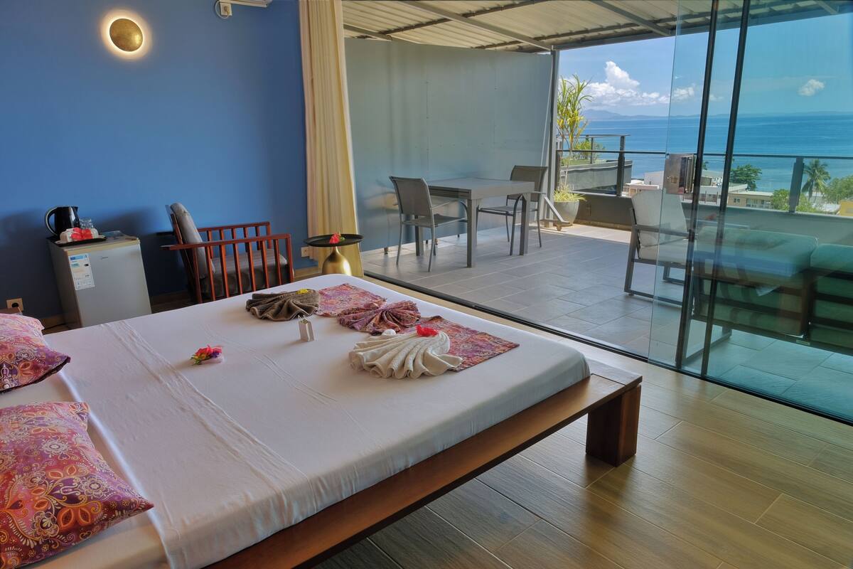 Habitación de hotel con cama, un pequeño escritorio, una silla, una mesa y un balcón con vistas al océano.