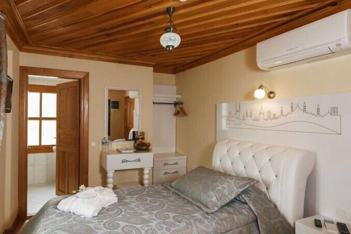 A bedroom with a wooden ceiling, a bed with patterned bedding, a bedside table, a mirror, and a wall-mounted air conditioner.