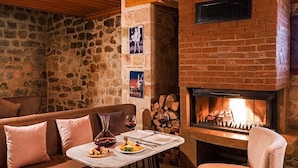 Um quarto aconchegante com parede de pedra, lareira de tijolos, uma mesa redonda com comida e assentos confortáveis.