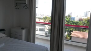 A bedroom with a bed, a bedside table, and a window with a view of buildings and trees. - Hotel Pousada Salvador (Salvador)