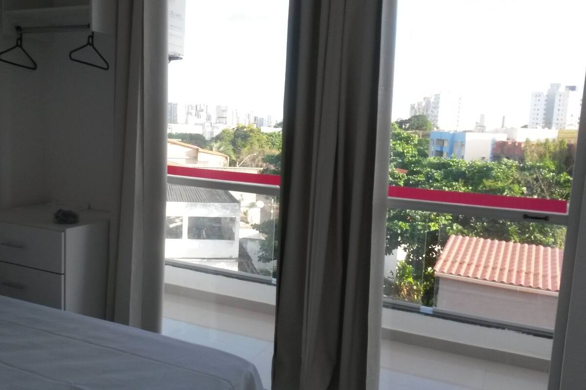 Une chambre à coucher avec un lit, une table de chevet et une fenêtre donnant sur des immeubles et des arbres.