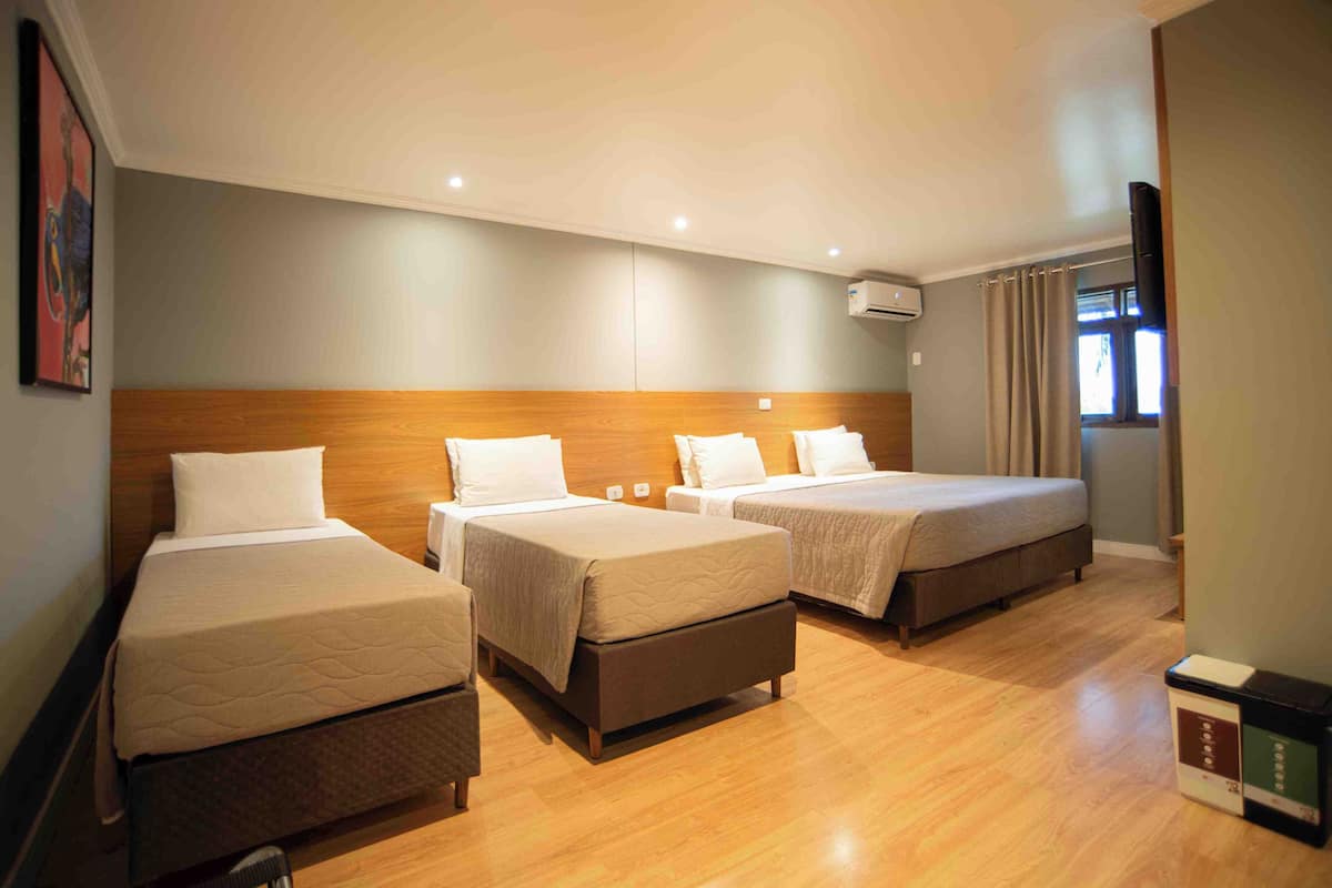 A hotel room with three beds, wooden headboard, a flat-screen TV, and an air conditioning unit.