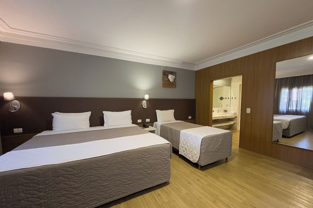 Hotellihuone, jossa on suuri sänky, kaksi pienempää sänkyä, puulattia, peili ja kylpyhuone, joka näkyy avoimesta ovesta.