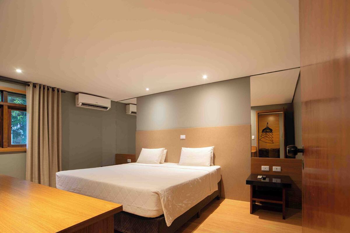 A hotel room with a bed, bedside table, air conditioning unit, and a mirror.