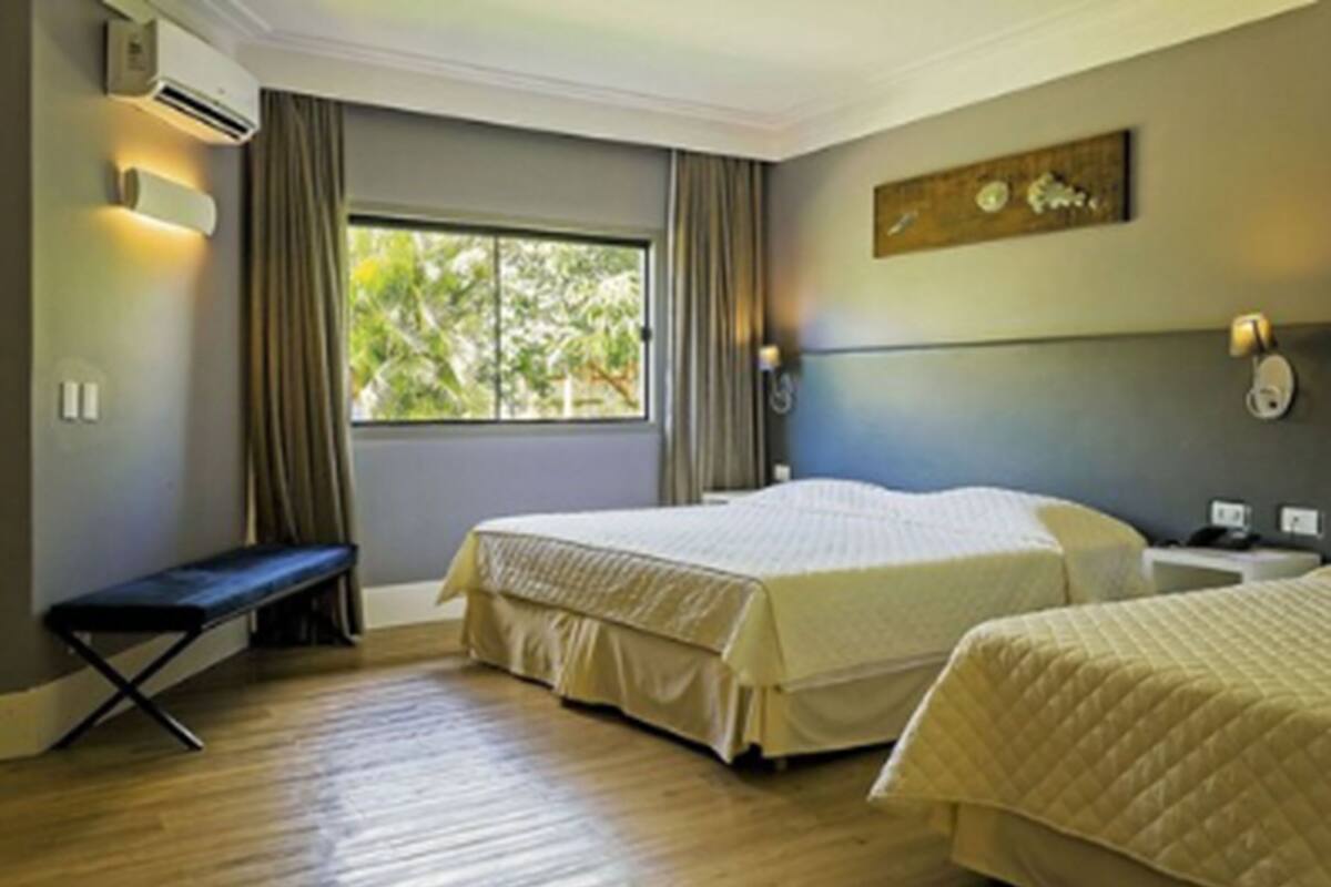 A hotel room with two beds, a bench, an air conditioning unit, a window with curtains, and a wall-mounted light fixture.