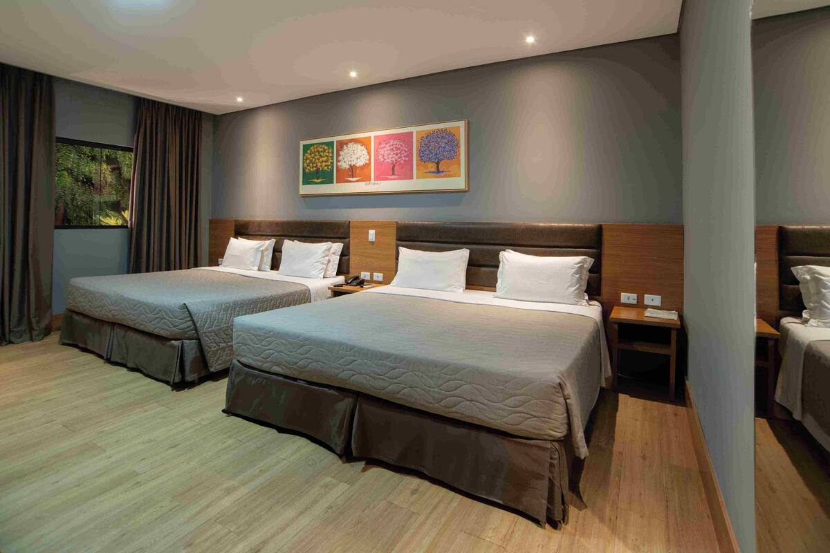 A hotel room with two beds, a large window with curtains, a painting on the wall, and a wooden floor.