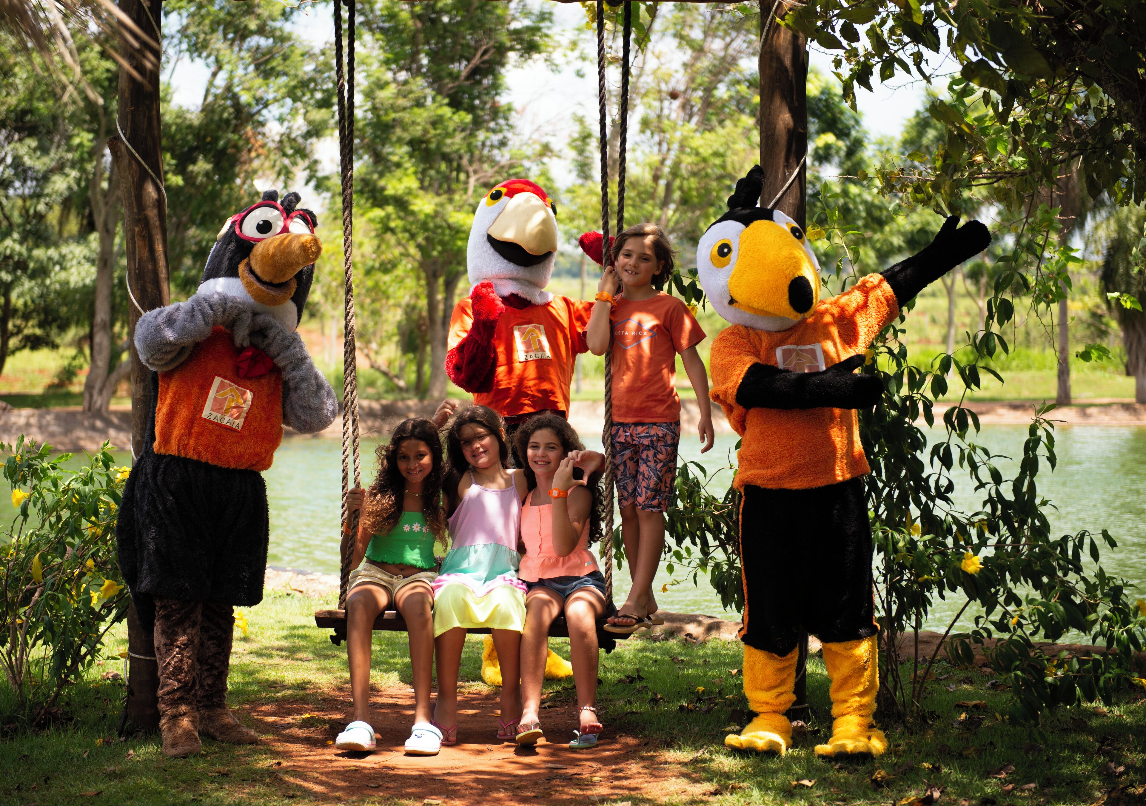 A group of children and adults interacting with large, colorful mascots in a natural setting with trees and a body of water.
