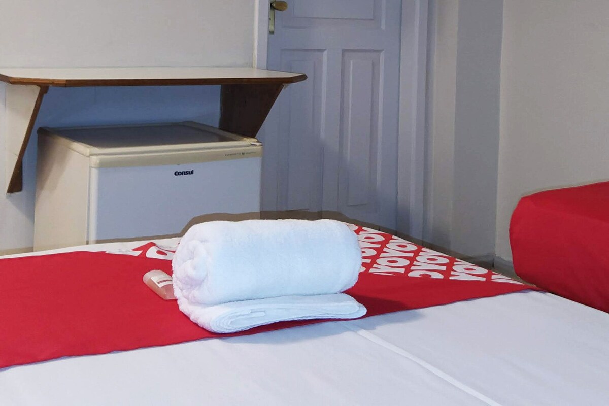 A hotel room with a bed, a TV mounted on the wall, a wooden shelf, and a small desk with a computer.