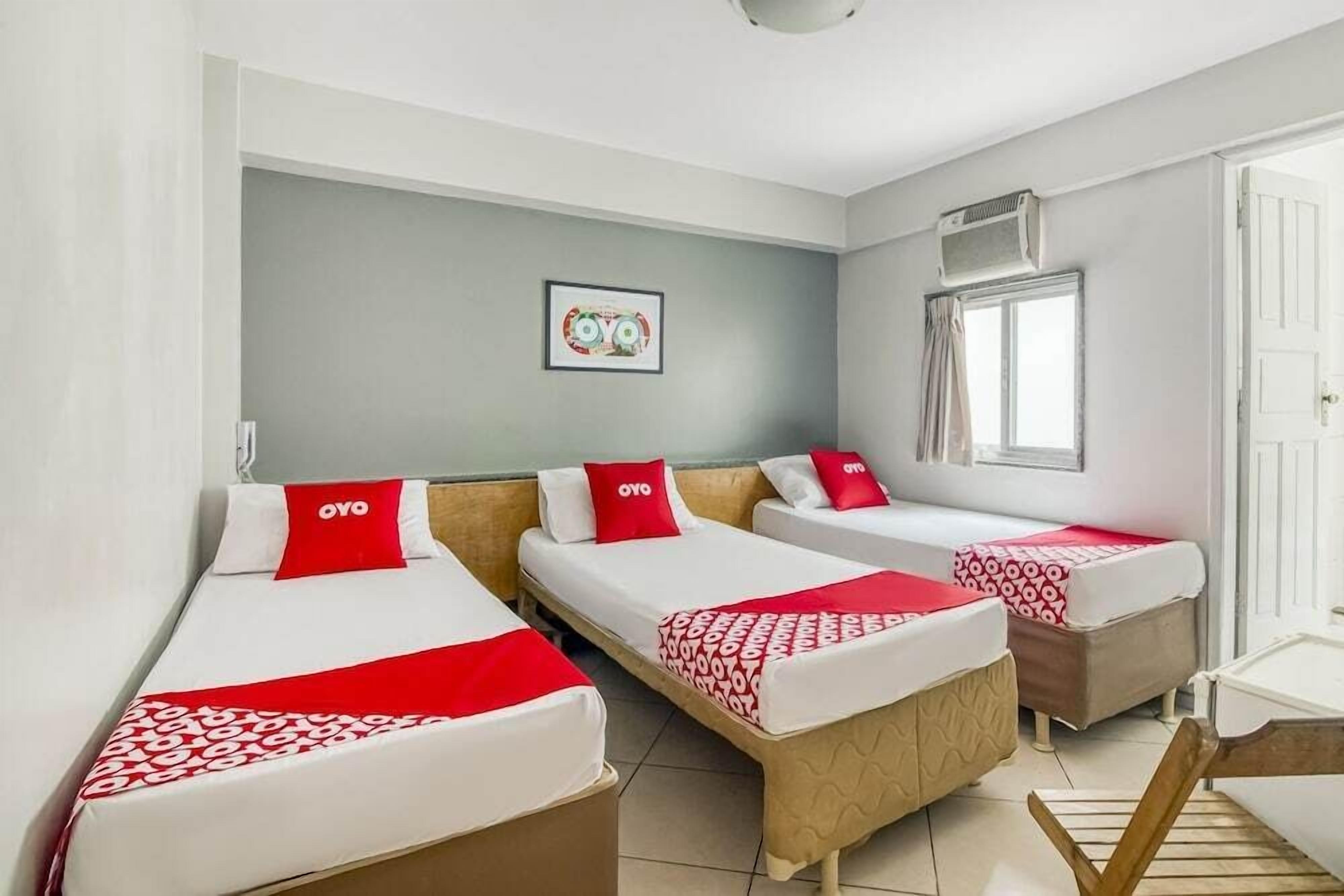 Quarto de hotel com três camas de solteiro, cada uma com um cobertor e travesseiro vermelhos, e um quadro na parede.