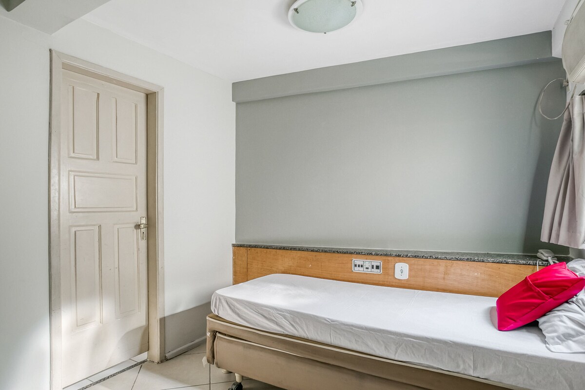 A single bed with a wooden headboard, a white mattress, and a red pillow. There is a door to the left and a light switch on the wall above the bed.