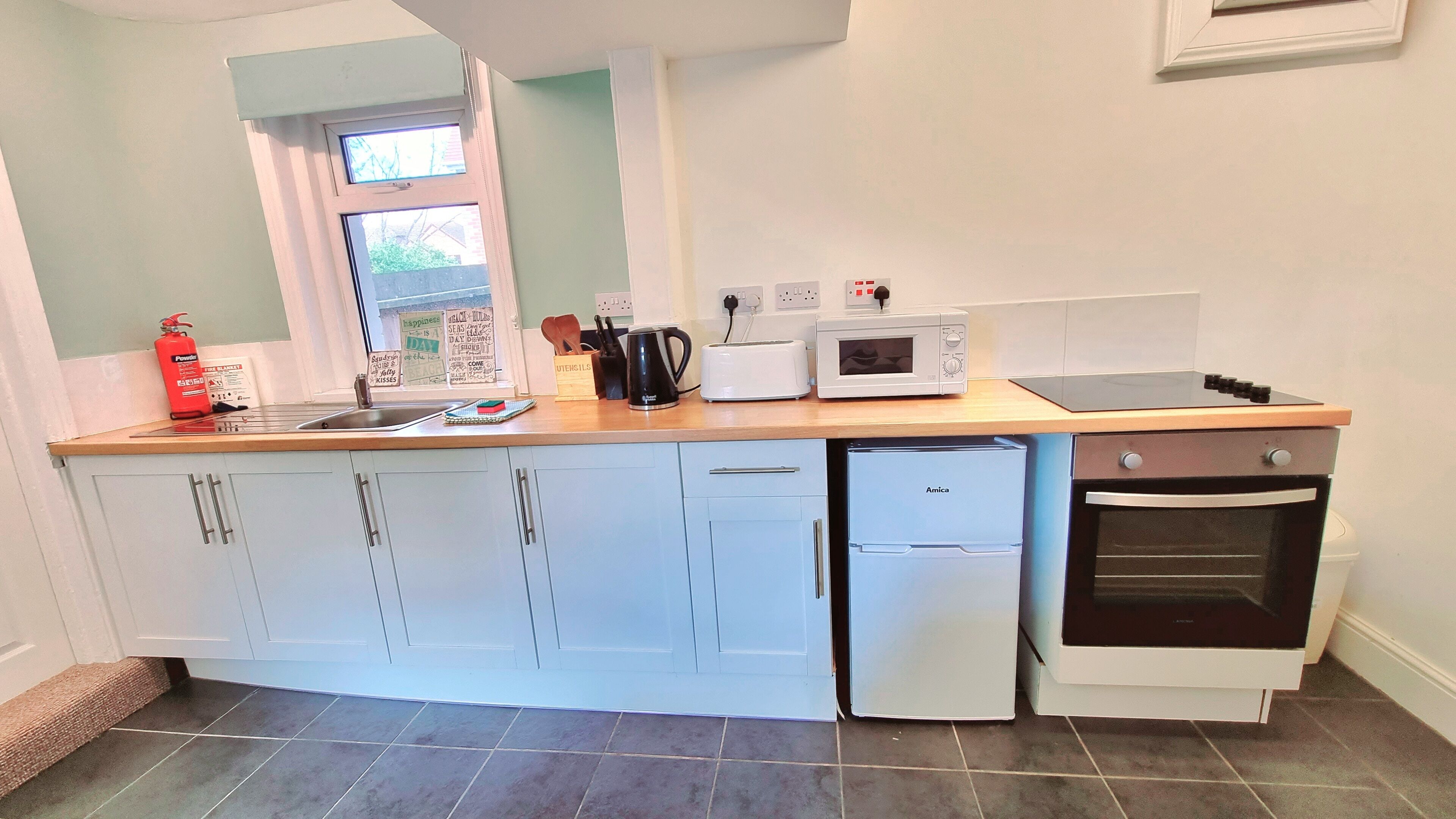 A compact kitchen with a dishwasher, oven, toaster, kettle, and a window with motivational posters.