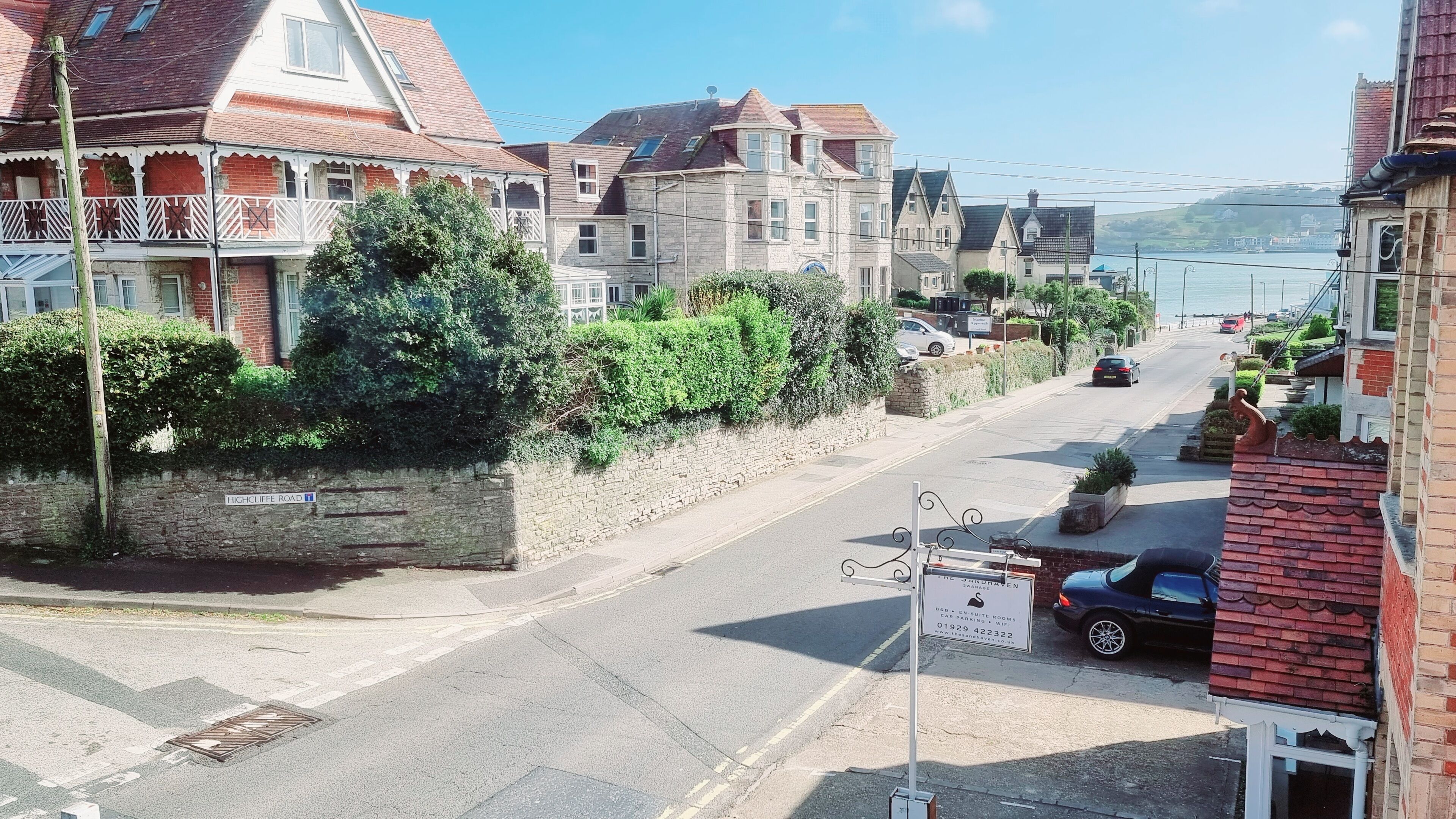 A street view with a row of traditional houses, a stone wall, and a clear view of the sea in the background.