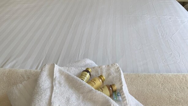 Cama bem arrumada com lençóis brancos e cabeceira escura com detalhe em matelassê. Uma toalha branca com produtos de higiene está sobre a cama.