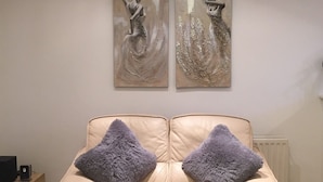 Um sofá de couro bege com duas almofadas fofas, uma mesa lateral de madeira com um alto-falante e duas pinturas abstratas na parede.