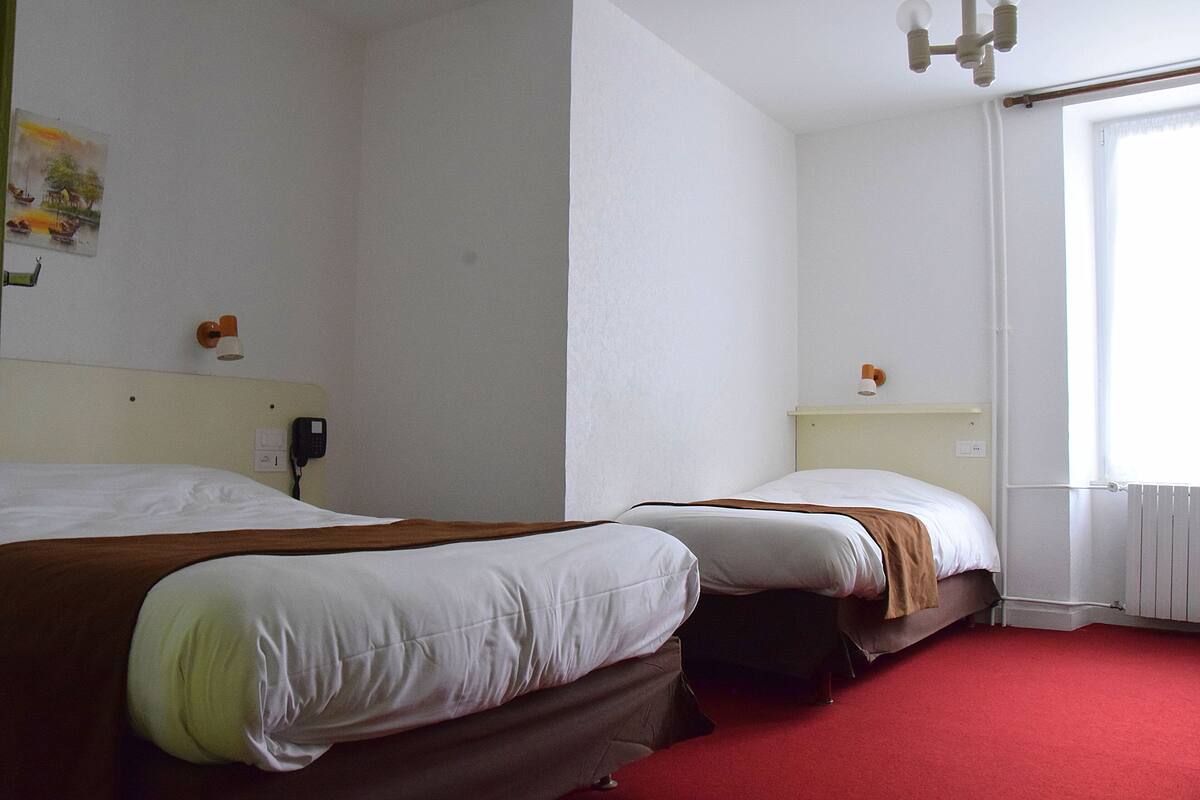 Habitación de hotel con dos camas, alfombra roja, pared blanca y ventana con cortinas.