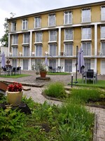 Un edificio amarillo con varios balcones, terraza al aire libre y un jardín con plantas en macetas.