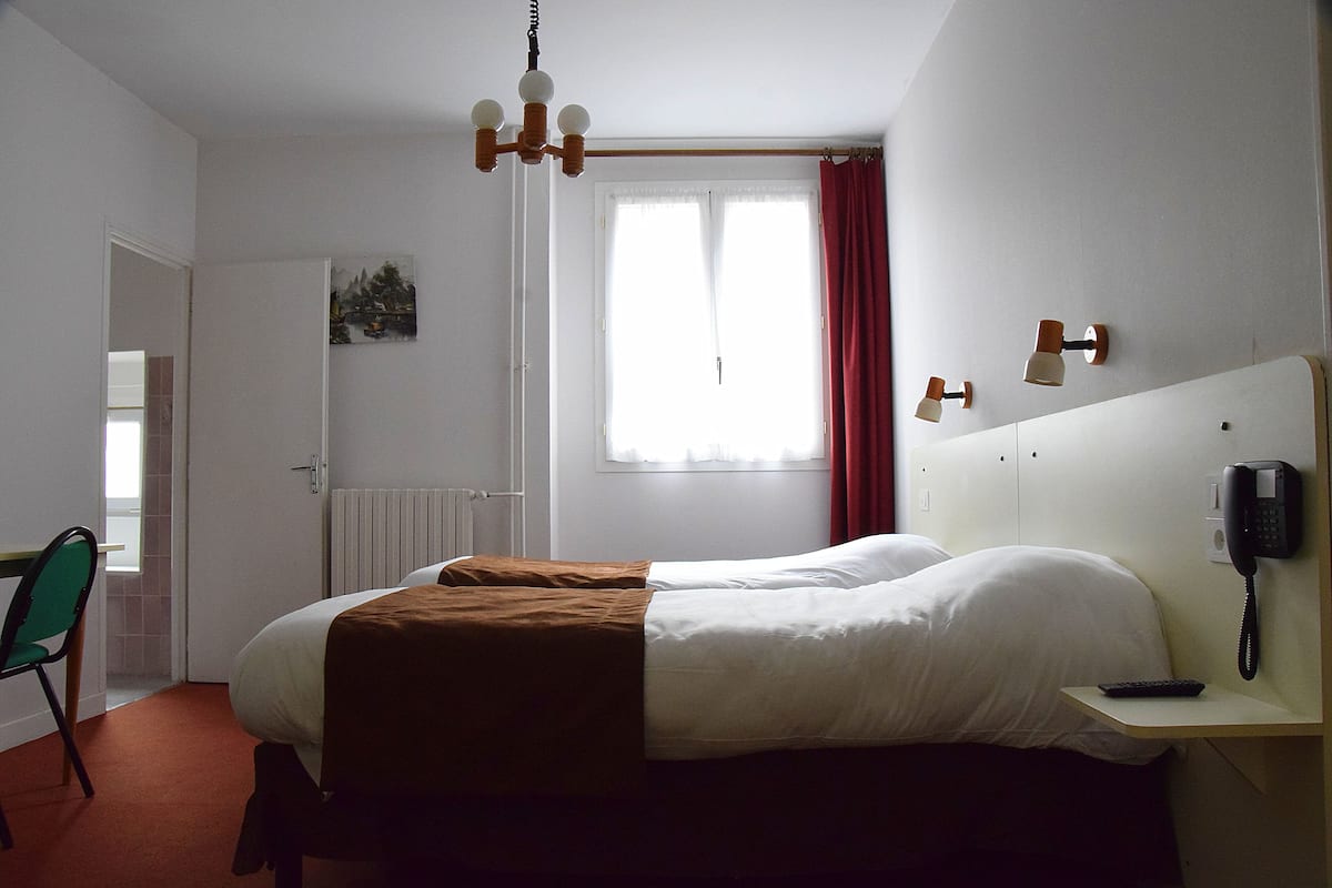 Habitación de hotel con cama, escritorio con teléfono, silla, radiador y ventana con cortinas.