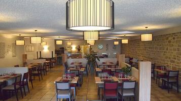 A restaurant with multiple dining tables and chairs, a central hanging light fixture, and a stone wall on one side.