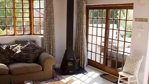 A cozy living room with a sofa, a coffee table with magazines, a fireplace, and large windows offering a view of the outdoors. - Sweetie Pie Clarens Self Catering Cottages (Clarens)