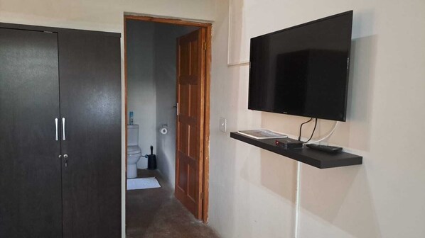 A hallway with a dark wooden wardrobe, a wooden door, a toilet, and a mounted flat-screen TV on a black shelf. - Iphofolo Lodge (Vivo)