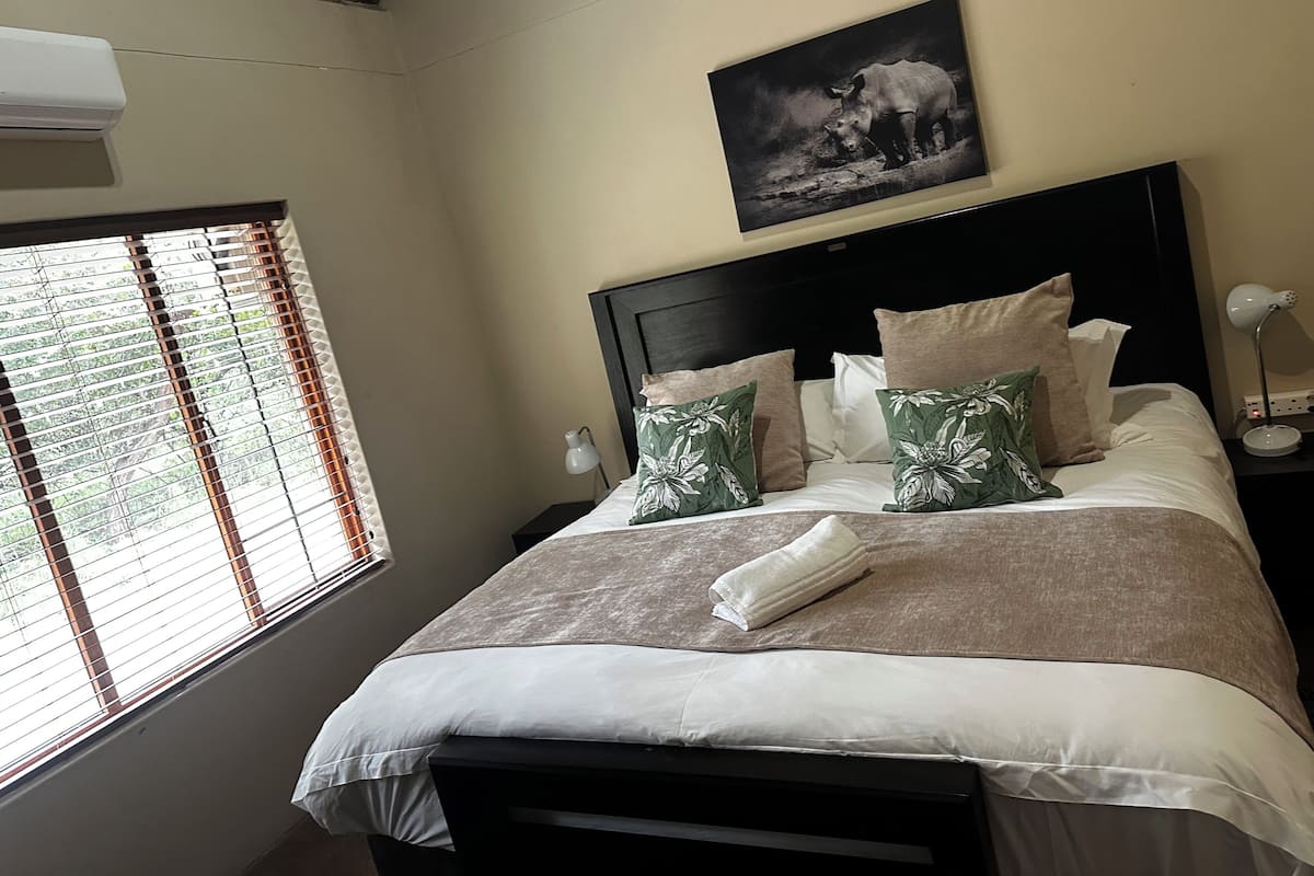 A neatly made bed with a white and brown bedspread, green floral pillows, and a framed picture of a rhinoceros on the wall.