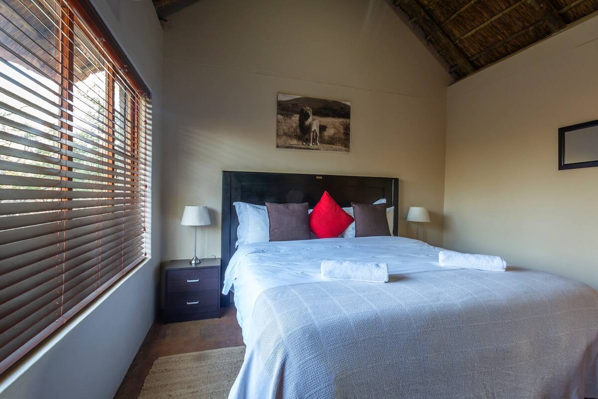 A bedroom with a bed, nightstands, a lamp, a painting on the wall, and a window with blinds.