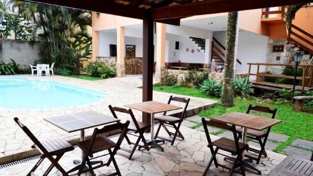 An outdoor patio with stone flooring, wooden tables and chairs, a pool, and surrounding greenery. - Pousada Conchas de Paraty (Paraty)