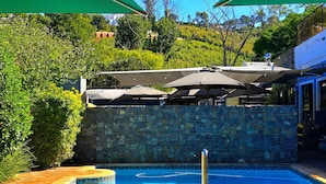 A pool area with a stone wall, two umbrellas, and lounge chairs. - Silver Forest Boutique Hotel, Spa, Restaurant & Bar (Cape Town)