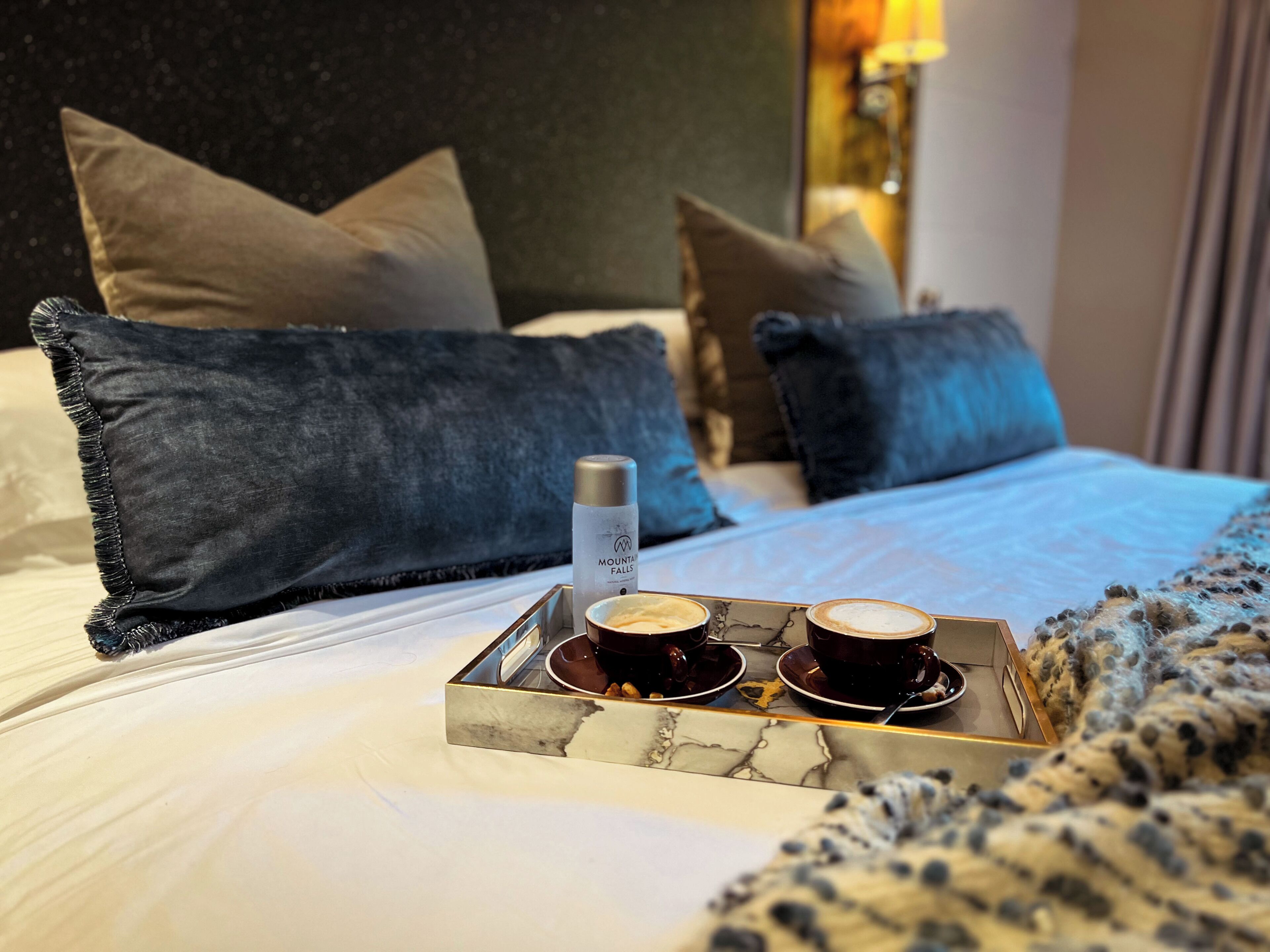 A neatly made bed with a marble tray holding two cups of coffee and a bottle of Mountain Falls water.