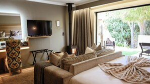 A hotel room with a sofa, a flat-screen TV, a fireplace, and a large window with a view of greenery. - Silver Forest Boutique Hotel, Spa, Restaurant & Bar (Cape Town)
