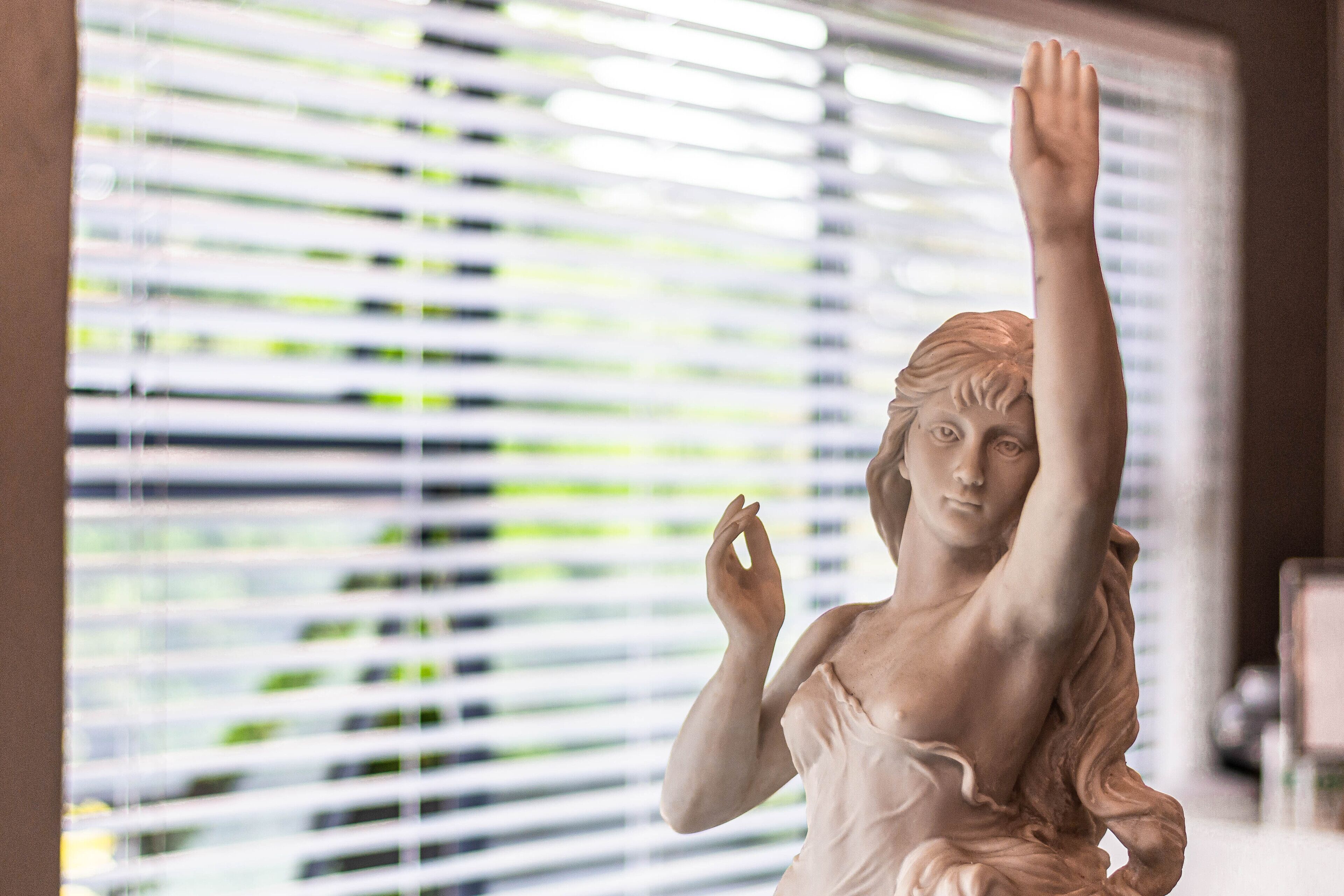 A statue with raised arms in a room with blinds in the background.