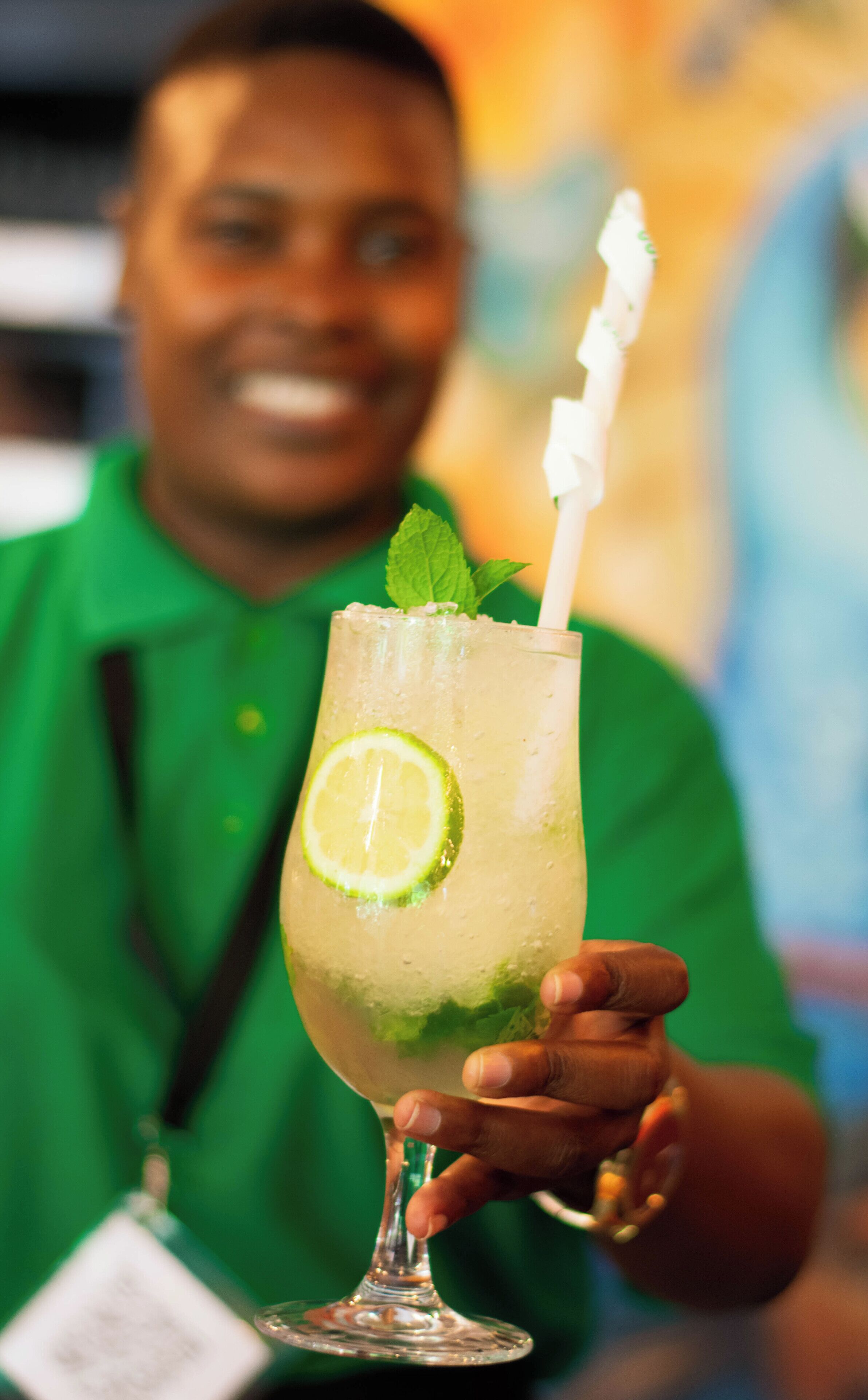 Eine Person hält ein Glas mit einer Limettenscheibe und Minze, sowie einen Strohhalm.