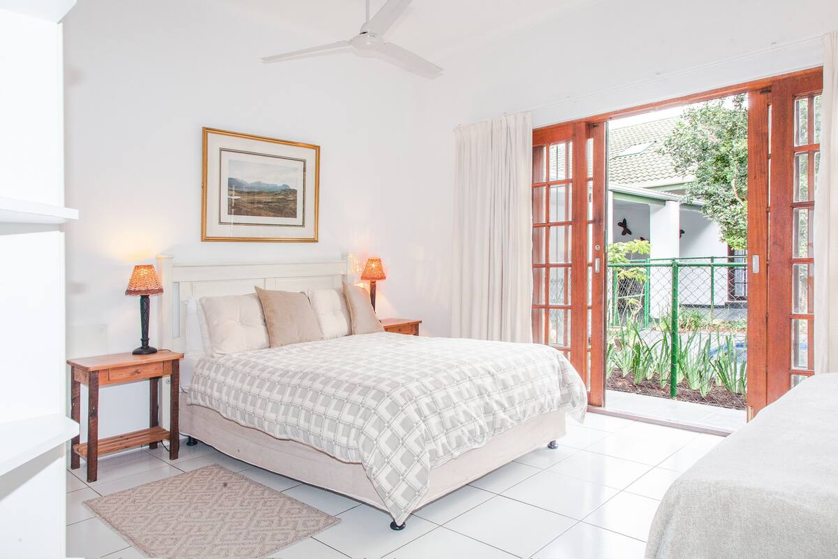 A bedroom with a bed, two bedside tables, a ceiling fan, a framed picture on the wall, and a view through an open door to a balcony with plants.