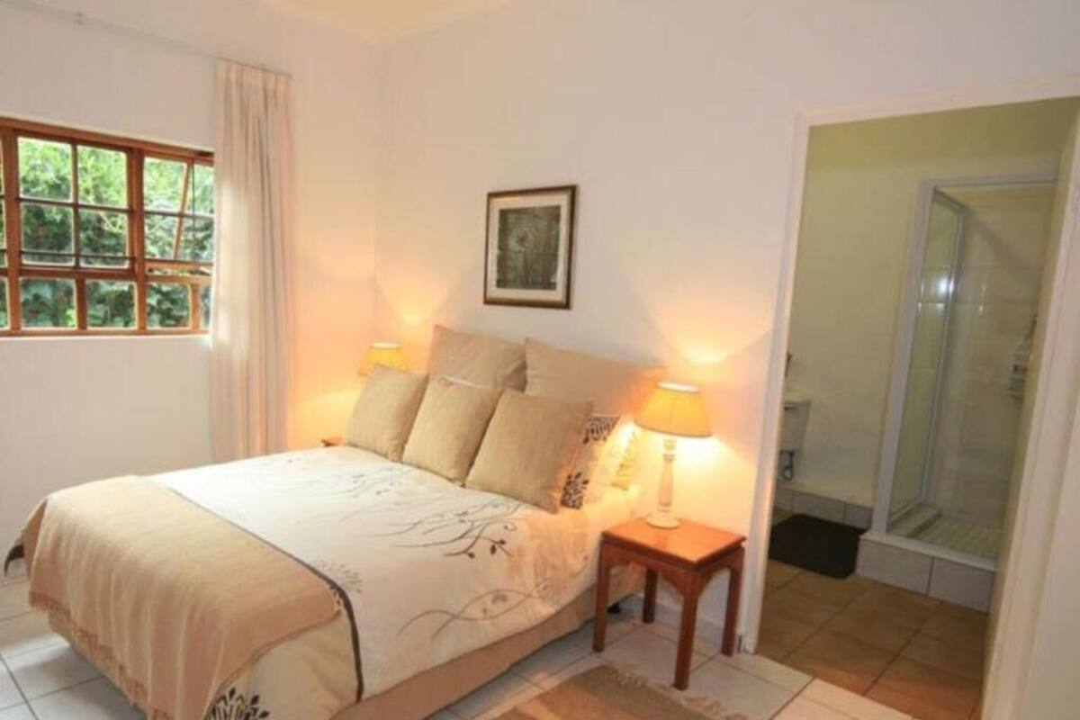 A bedroom with a bed, bedside lamps, a framed picture on the wall, a window, and a shower area.